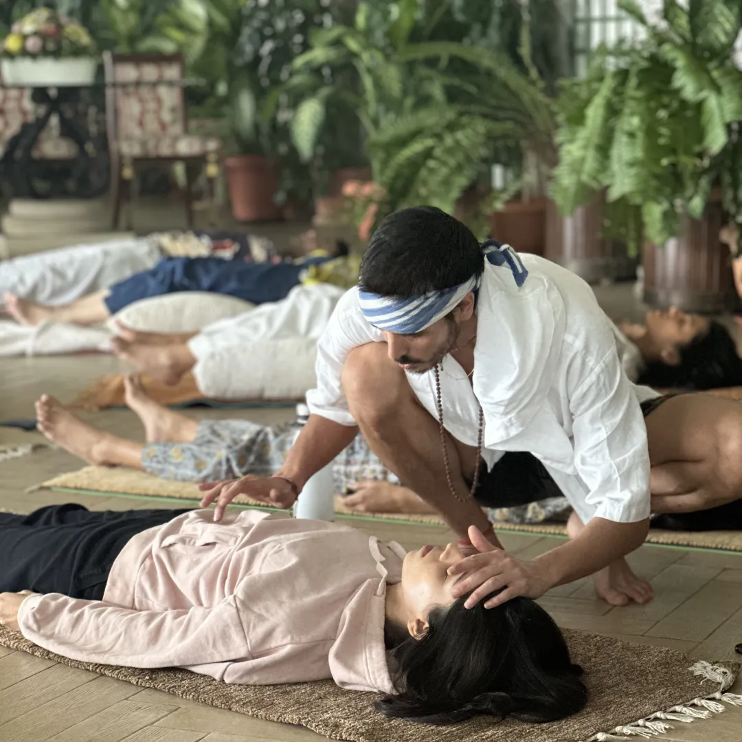 Elliot Amaru, a blue and white headband in his hair, leaning over a woman lying down, one hand on her forehead, during a group session