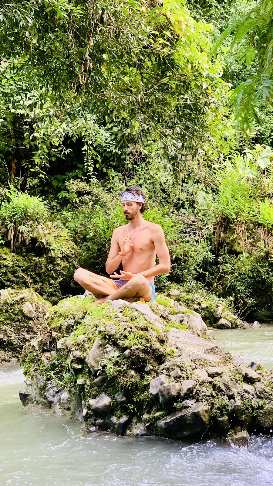 Elliot Amaru assis en lotus sur un rocher au milieu d'une rivière, entouré de forêt, la main sur le cœur