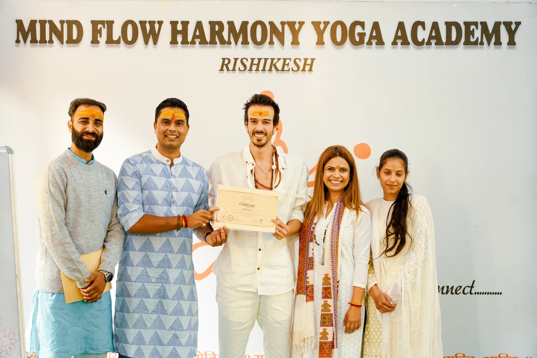 Elliot Amaru reçoit son certificat de yoga, entouré de ses professeurs, devant le panneau de la Mind Flow Harmony Yoga Academy à Rishikesh